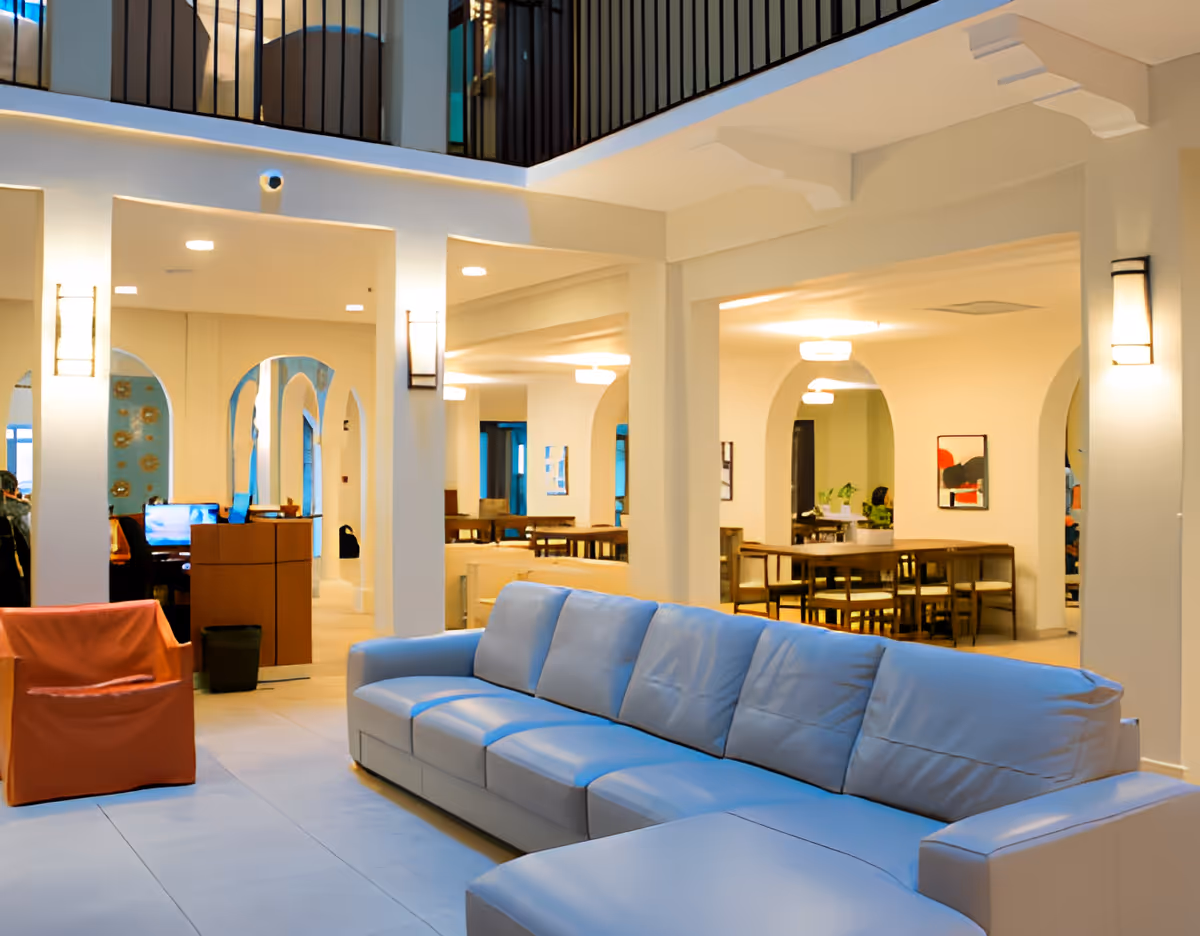 Spacious and well-lit interior of a senior living facility featuring a large light gray sectional sofa and an orange armchair in the foreground. The background shows a dining area with wooden tables and chairs, white walls with arches, and modern ceiling lights. The space has a clean, welcoming atmosphere with soft lighting and decorative wall sconces.