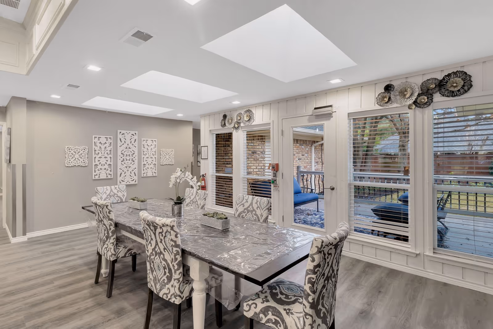 Bright dining area with patterned chairs around a long table, skylights overhead and large windows and a door opening to an outdoor patio.