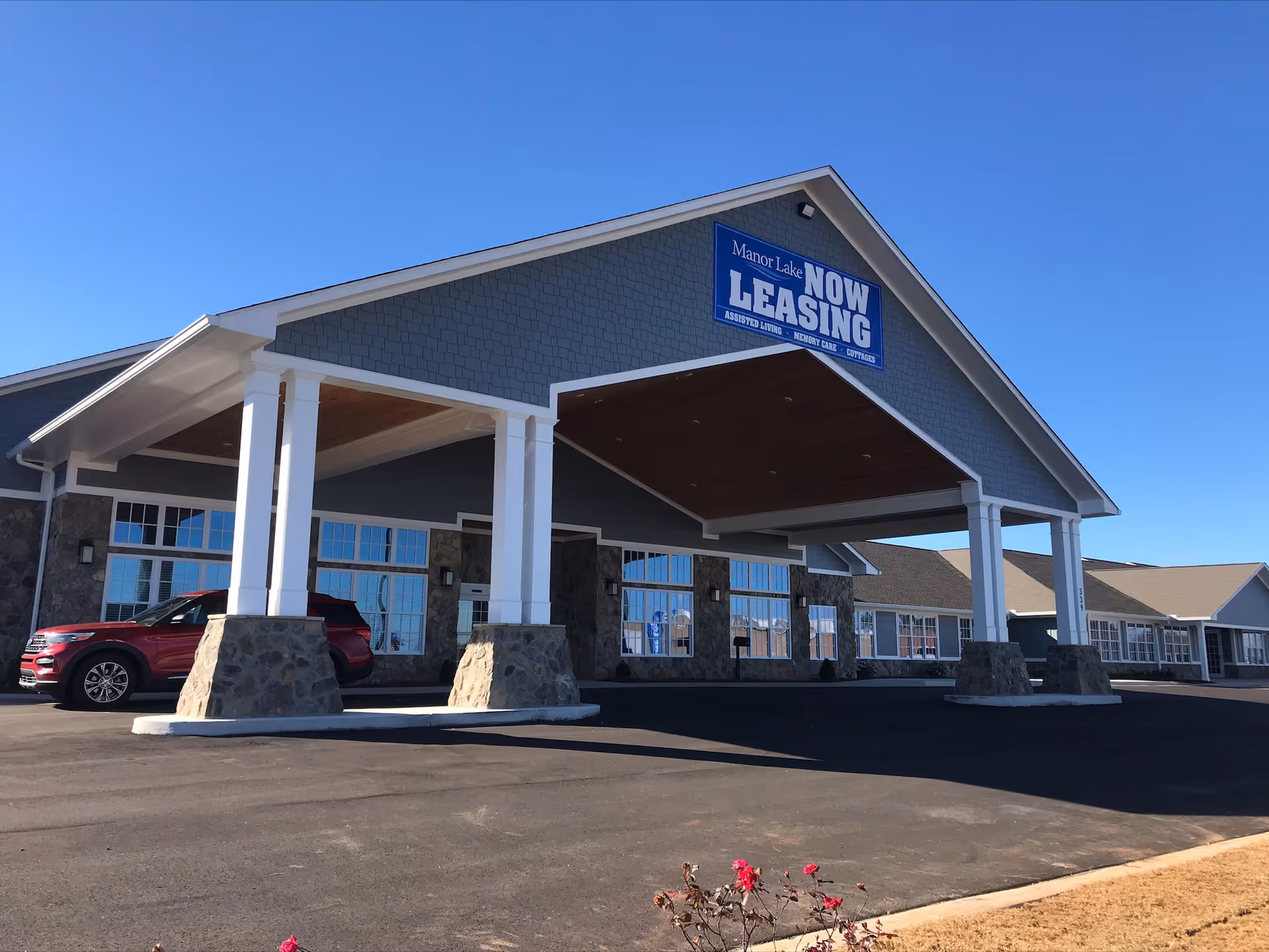 Exterior view of Manor Lake Assisted Living & Memory Care facility with a large covered entrance supported by white columns with stone bases. A red SUV is parked under the entrance. A blue sign on the building reads 'Manor Lake NOW LEASING Assisted Living Memory Care Courtyard'. The sky is clear and blue.