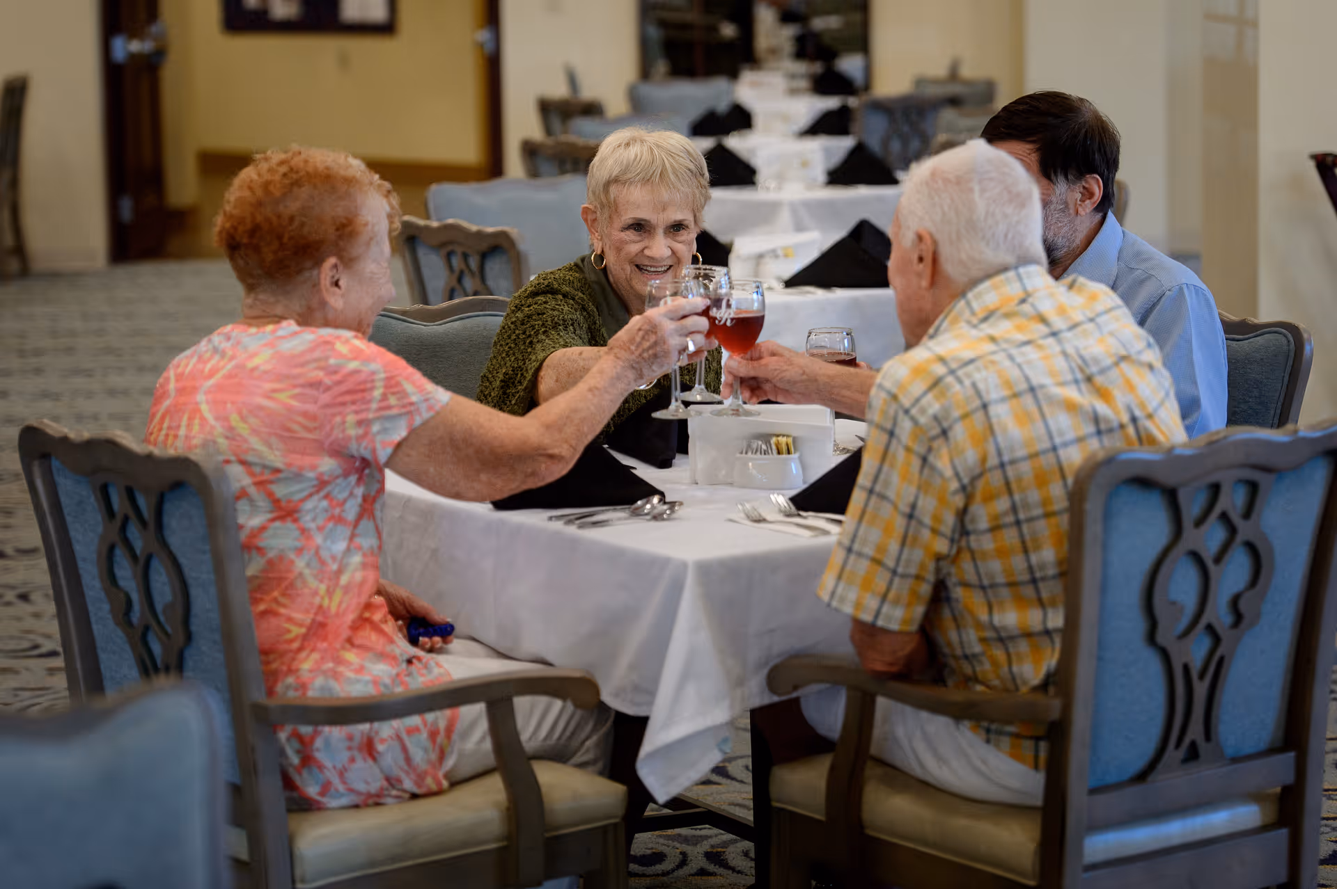 Four older adults seated at a dining table clinking glasses in a communal dining room.