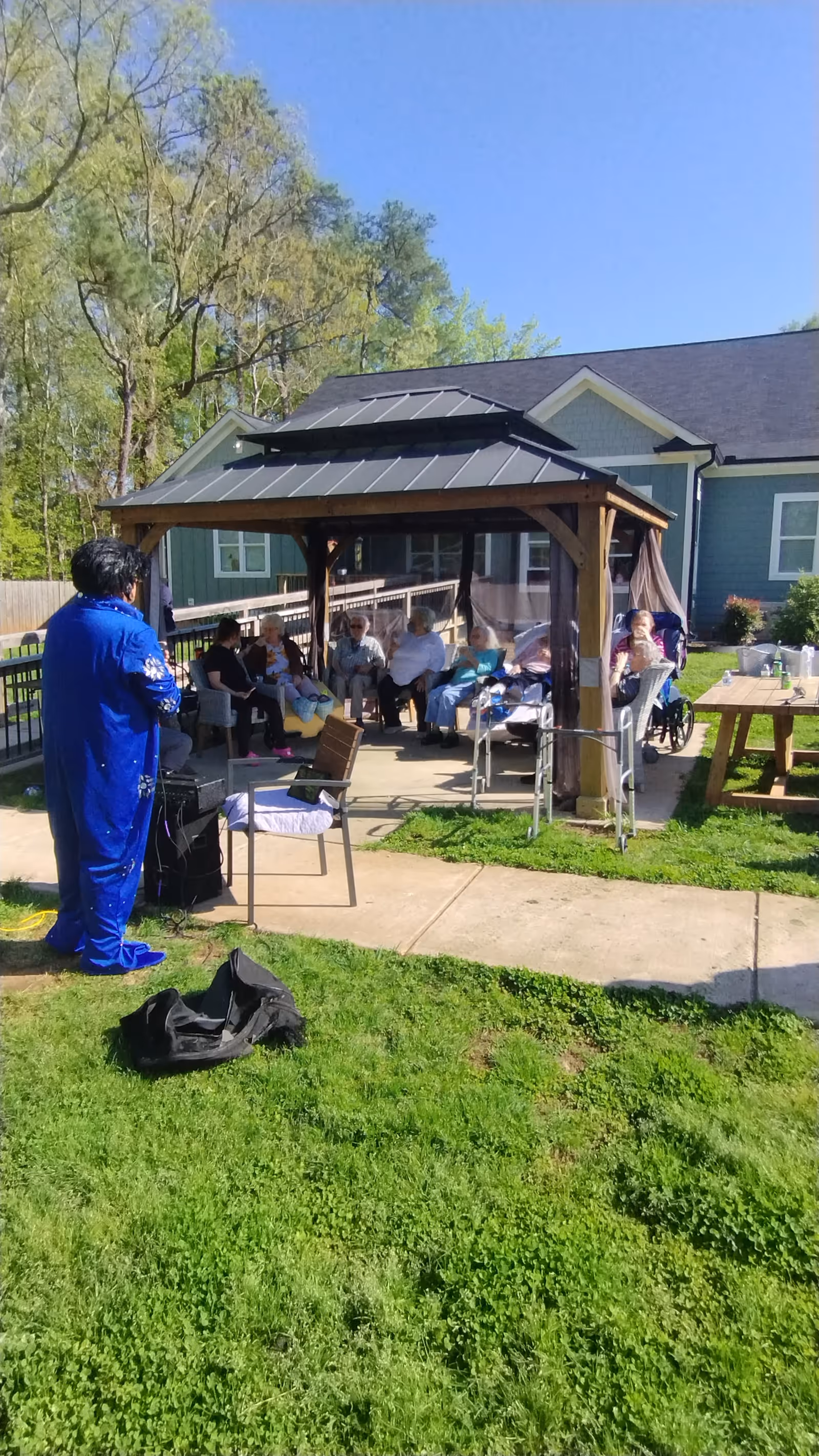 A performer in blue entertains a group of seated senior residents under a wooden gazebo on the lawn outside a green-sided building.