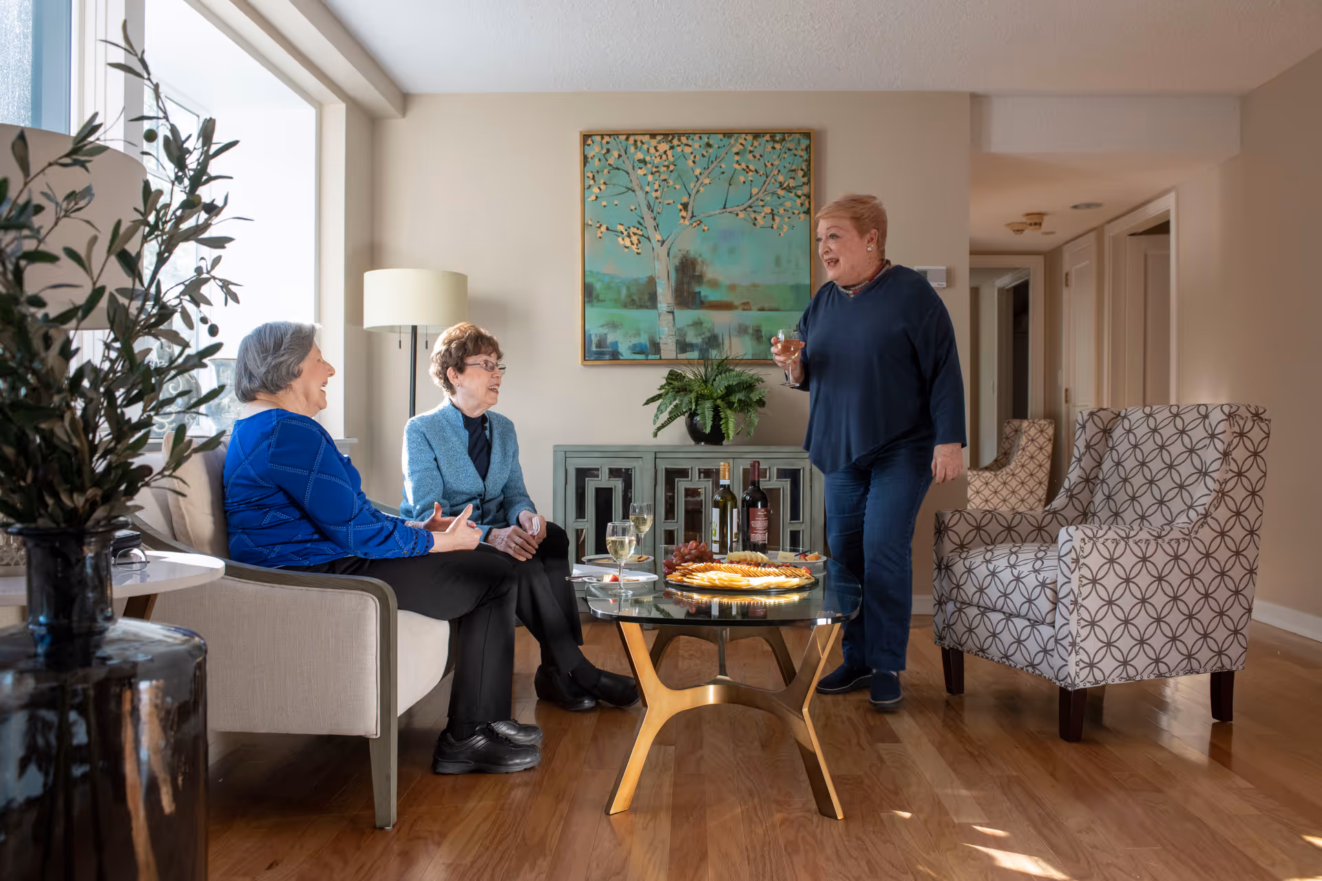 Three elderly women socializing in a well-lit living room. Two women are seated on a beige couch, and one woman is standing holding a glass of wine. A glass coffee table in front of them holds a plate of snacks, two glasses of white wine, and two bottles of wine. The room has wooden flooring, a large window, a floor lamp, a decorative plant, and a painting of a tree on the wall.