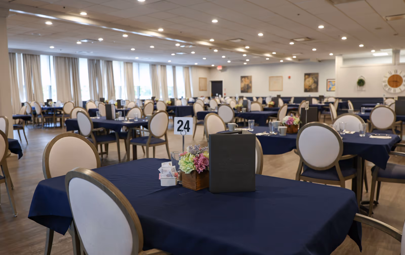 A spacious dining room with multiple tables covered in dark blue tablecloths. Each table has beige cushioned chairs, a small floral centerpiece, a menu, and table number holders. The room is well-lit with ceiling lights and has large windows with beige curtains along one side.