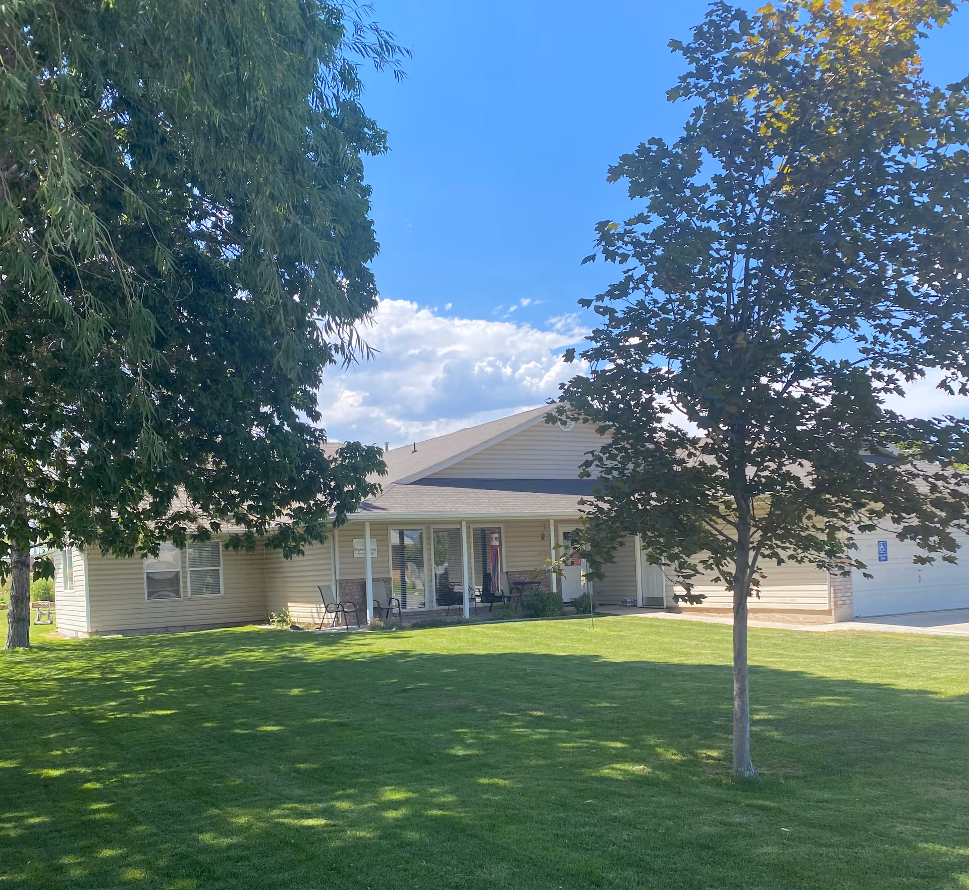 Exterior view of a single-story beige building with a covered porch, surrounded by green grass and trees under a blue sky with some clouds.