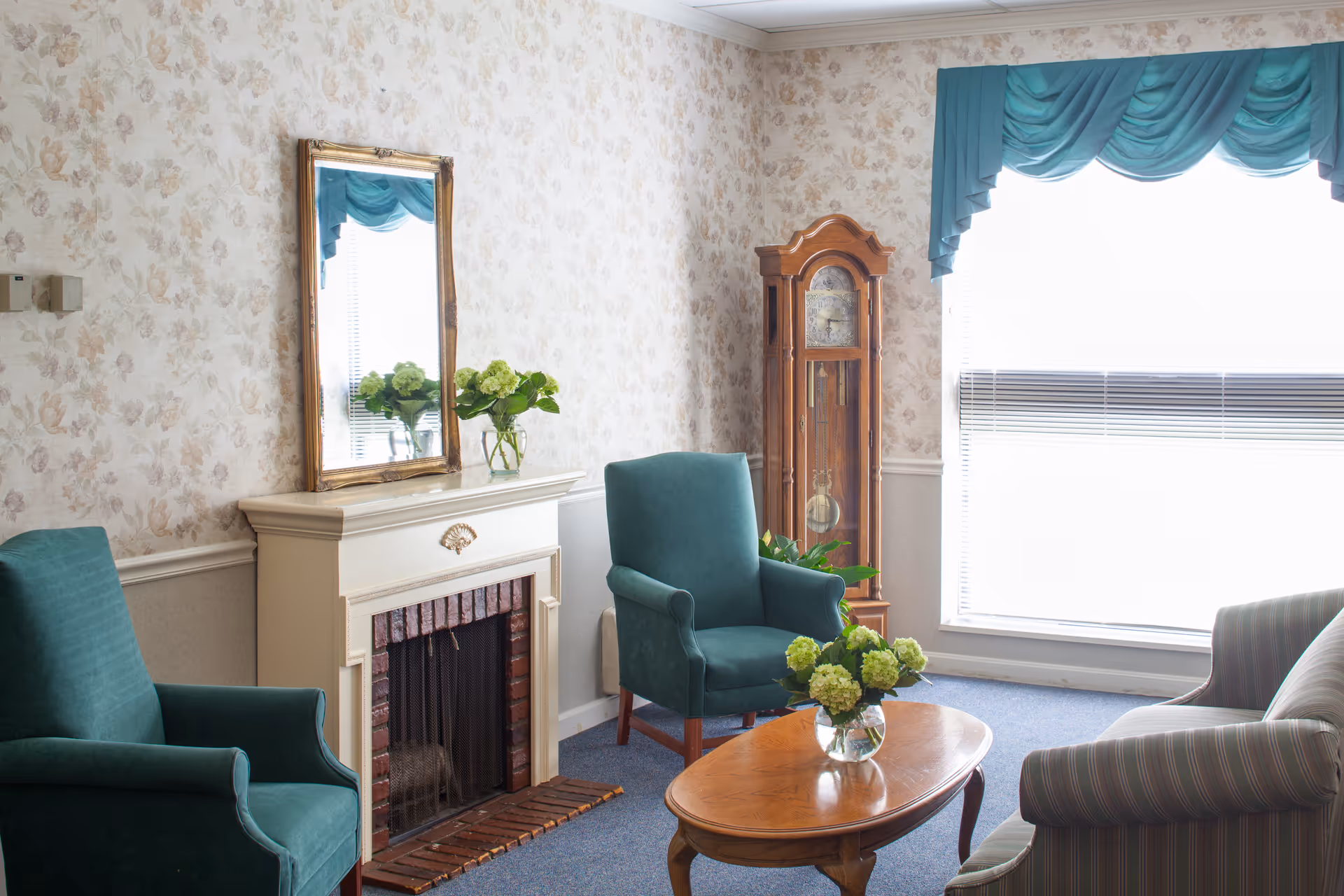 A cozy living room with floral wallpaper, a white fireplace with a mirror above it, two teal armchairs, a striped sofa, a wooden coffee table with a vase of green flowers, a tall wooden grandfather clock, and a large window with teal curtains.