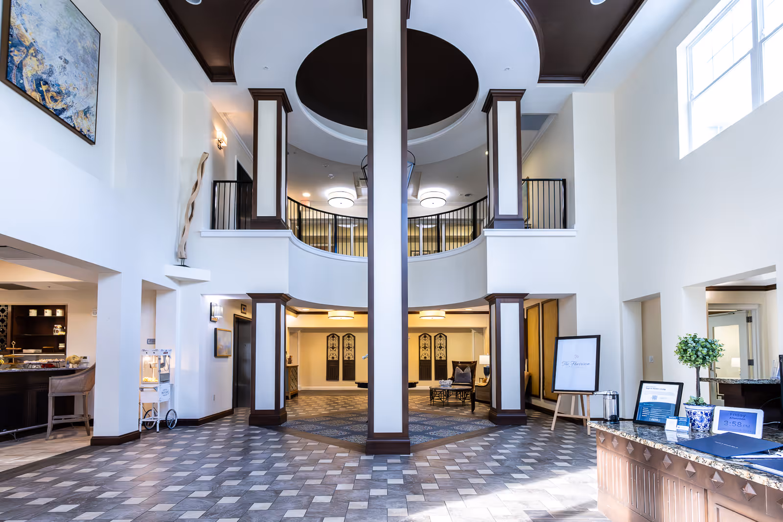 Spacious and well-lit interior lobby area of The Harrison of Wildwood featuring high ceilings, large windows, decorative columns, and a patterned tile floor. The space includes seating areas, a reception desk with informational materials, and modern light fixtures. Artwork and plants add to the welcoming atmosphere.