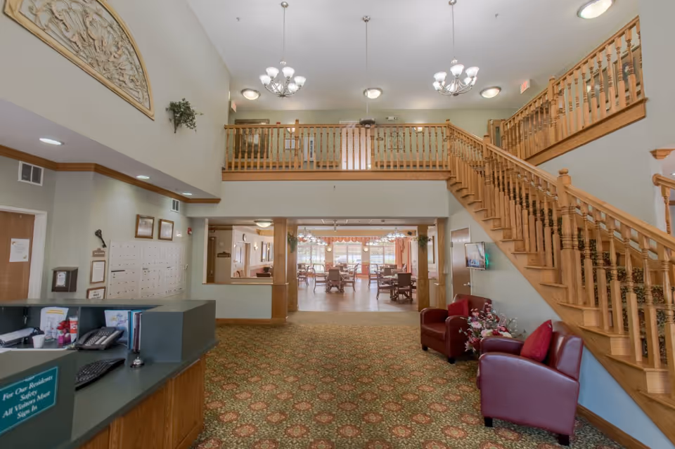 Interior view of a senior living facility lobby area with a reception desk on the left, two red armchairs with a floral arrangement on the right, and a wooden staircase leading to an upper floor. Beyond the lobby is a dining area with tables and chairs, illuminated by chandeliers and natural light from large windows.