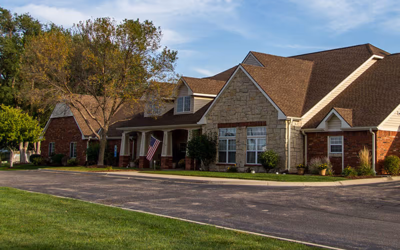 Exterior view of a senior living facility building with a combination of brick and stone facade, a brown shingled roof, several windows, and a covered entrance with an American flag. The building is surrounded by trees, shrubs, and a paved driveway.