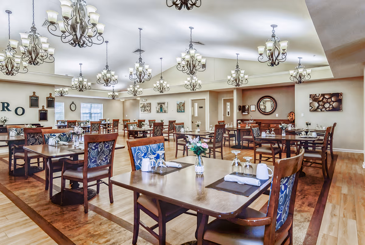 A spacious dining room with multiple wooden tables and chairs arranged neatly. Each table is set with placemats, napkins, glasses, and small flower vases. The room features elegant chandeliers hanging from the ceiling, light-colored walls, and wooden flooring. Decorative wall art and a round mirror are visible on the walls.
