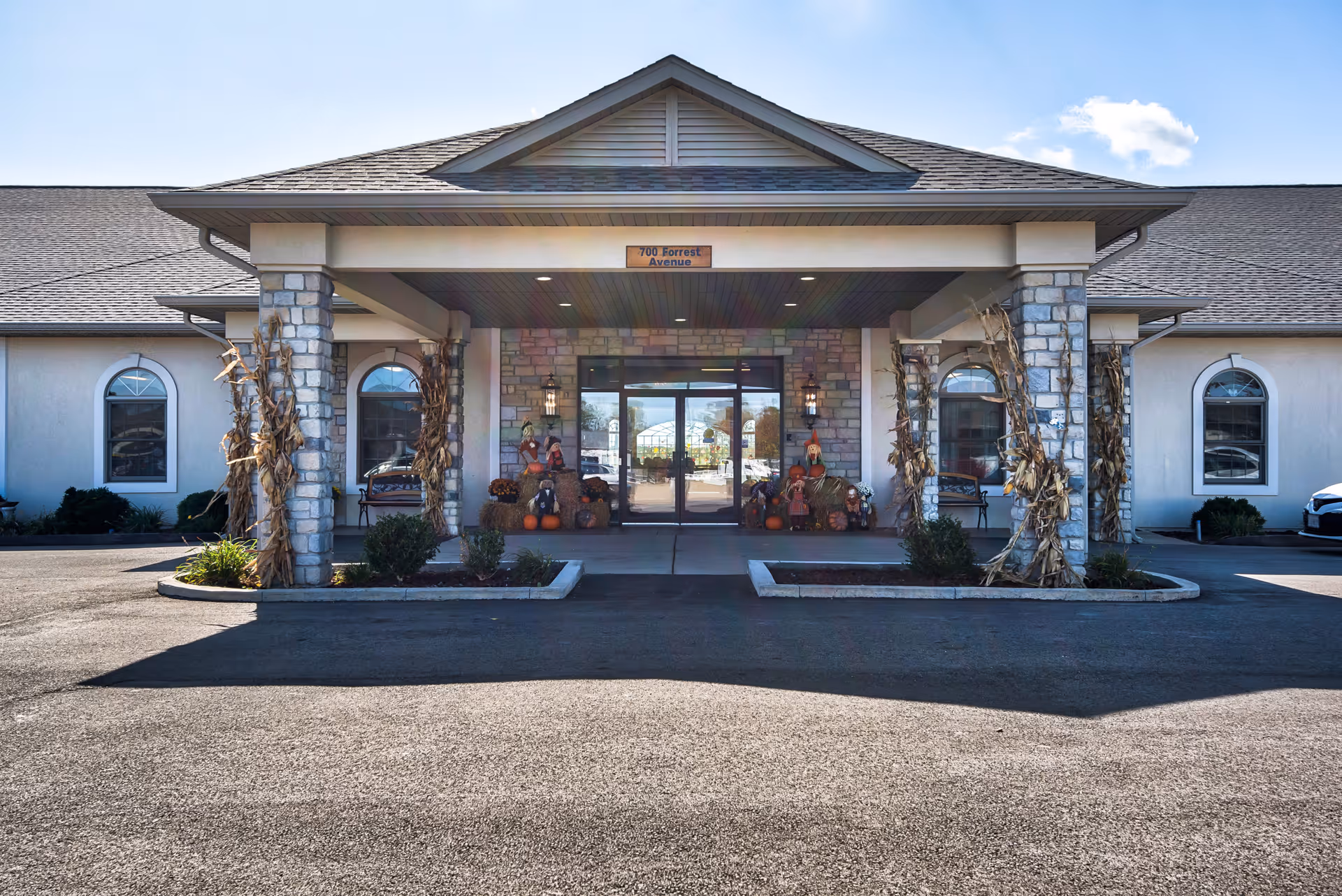 Covered front entrance of a single-story assisted living building with stone columns and autumn decorations including corn stalks and pumpkins.