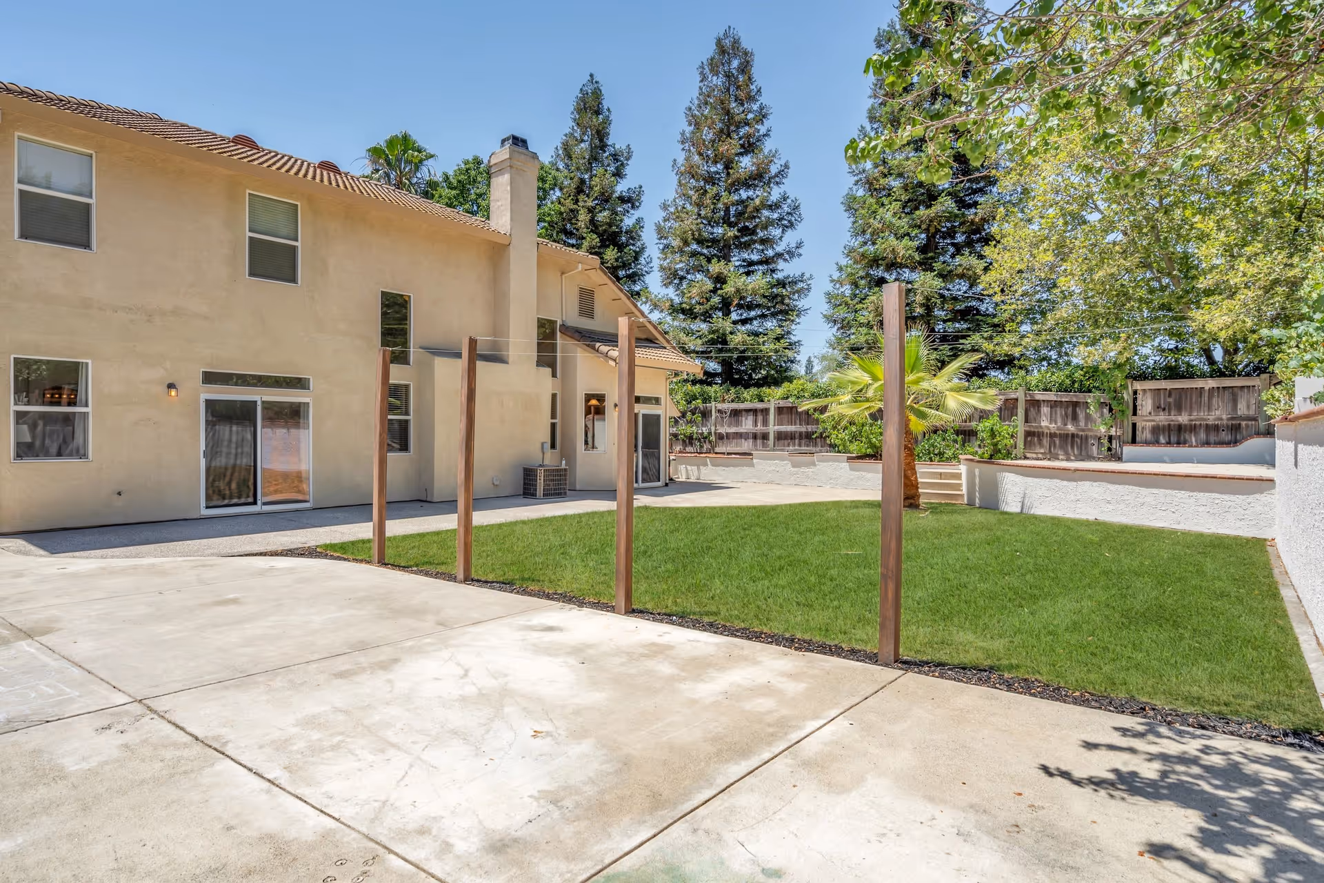 Backyard of a two-story residence with a lawn, concrete patio, wooden posts, and a surrounding fence.