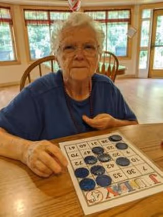 An elderly woman with white hair and glasses sitting at a wooden table playing bingo. She is placing black markers on a bingo card. The room has large windows with a view of greenery outside and wooden chairs around the table.
