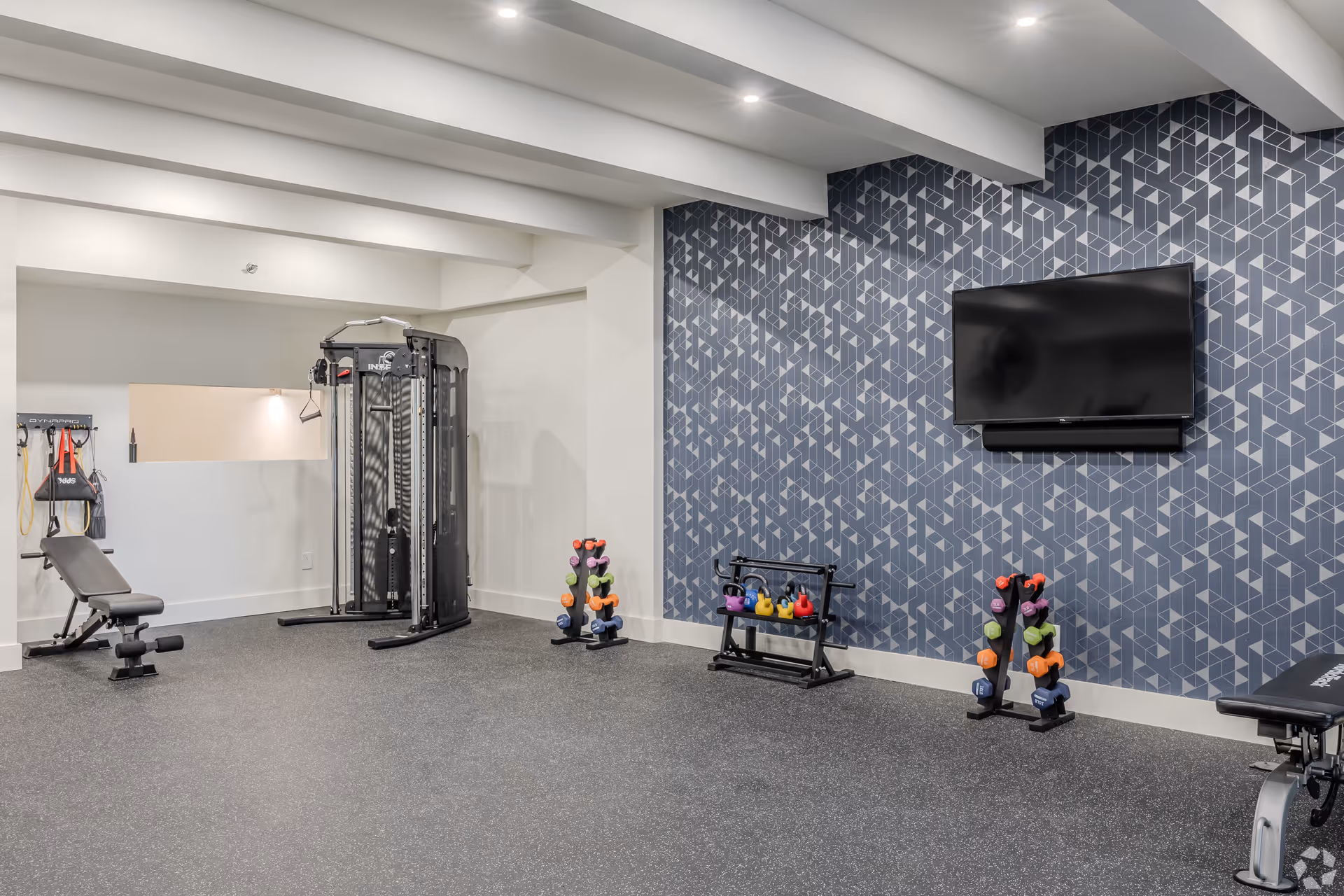 A modern fitness room with exercise equipment including a weight bench, a multi-functional cable machine, and racks holding colorful dumbbells and kettlebells. The room has a geometric patterned blue accent wall with a mounted flat-screen TV and recessed ceiling lights.