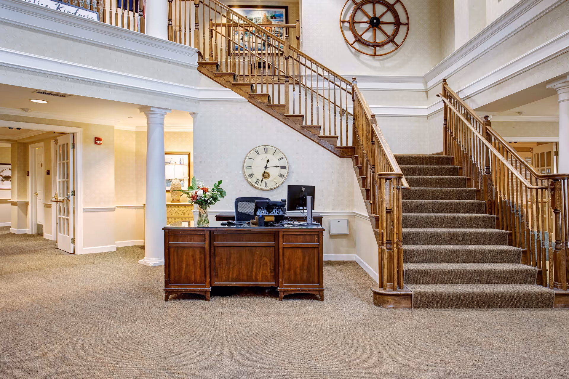 Interior view of a senior living facility lobby area with a wooden reception desk, a computer, a vase with flowers, a large wall clock, and a carpeted staircase with wooden railings leading to the upper floor. The walls are light-colored with decorative molding, and there are columns supporting the upper level.