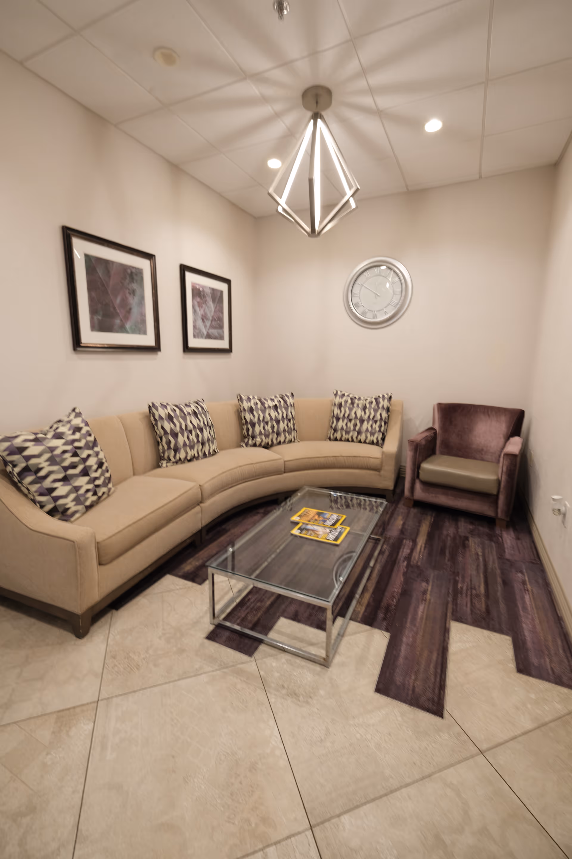 Small lounge with a curved beige sofa and patterned pillows, a glass coffee table, a purple armchair, framed art, a wall clock, and a geometric pendant light.