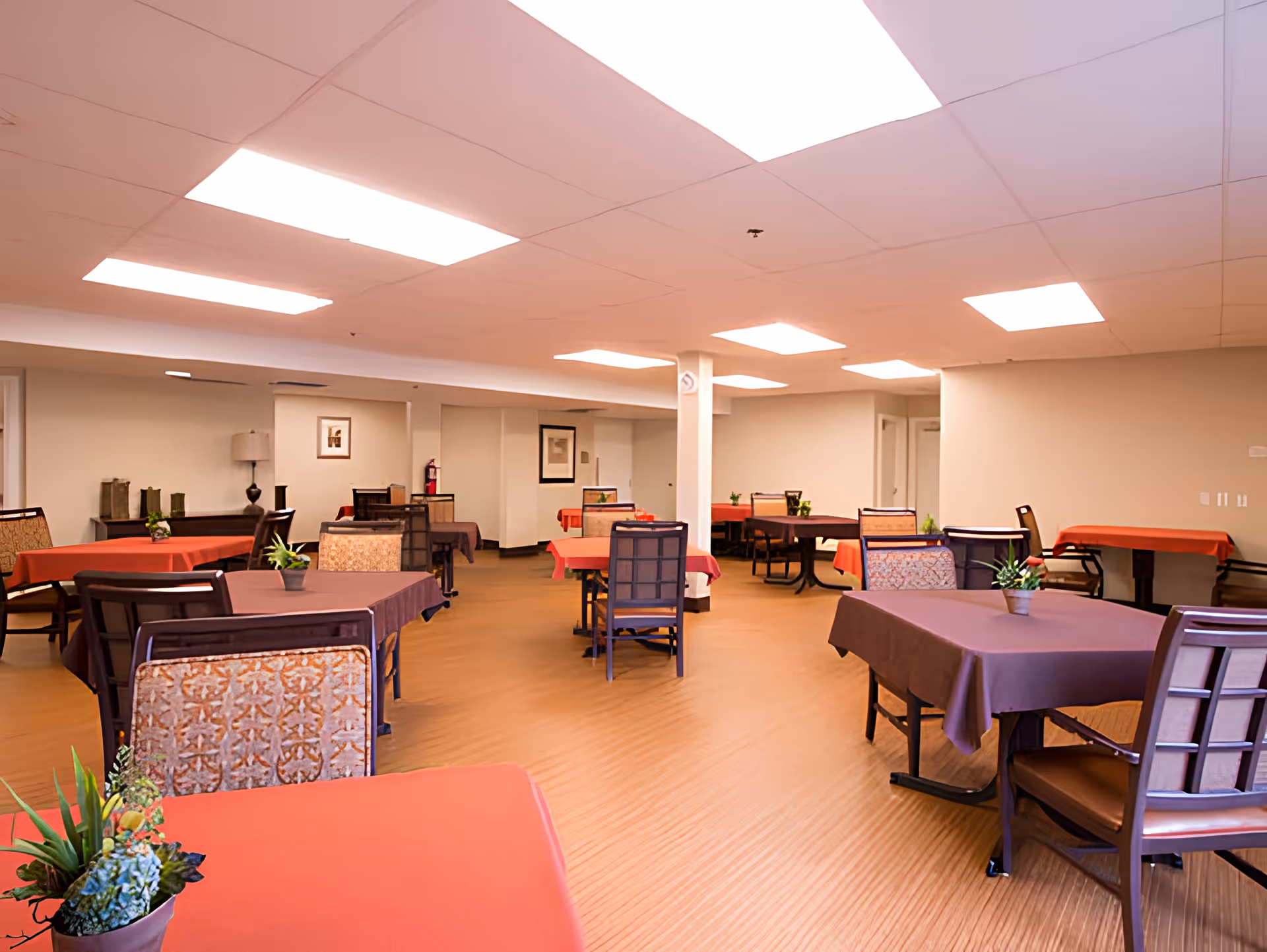 A spacious dining room with multiple tables covered in red and brown tablecloths, each adorned with small potted plants. The room has beige walls, a light brown floor, and bright overhead lighting. Chairs with patterned upholstery and wooden frames are arranged around the tables.
