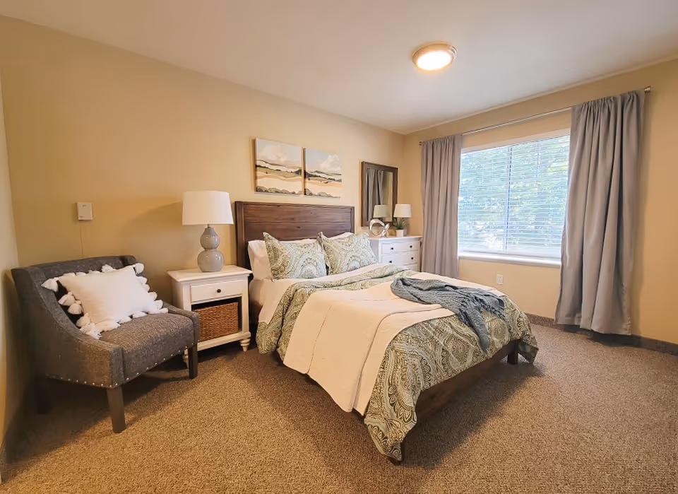 A cozy bedroom with a large window covered by gray curtains, a bed with patterned bedding and multiple pillows, a white nightstand with a lamp, a gray armchair with a white pillow, a white dresser with a mirror and another lamp, and beige carpeted floor.