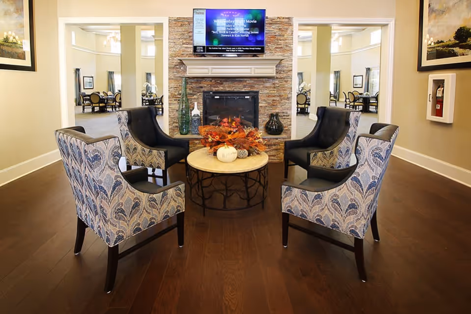 A cozy seating area with four patterned armchairs arranged around a round table with autumn-themed decorations, in front of a stone fireplace with a TV mounted above it. The room has wooden flooring and opens into a dining area with tables and chairs.