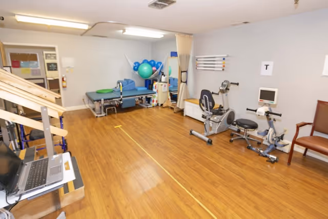 A physical therapy room with wooden flooring, exercise equipment including a stationary bike, therapy balls, a treatment table, and a laptop on a stand. The room has light gray walls and fluorescent ceiling lights.