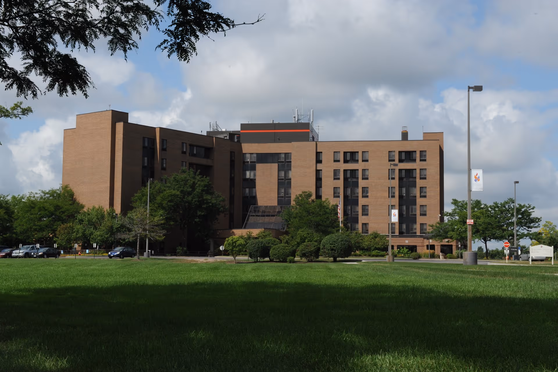 Exterior view of a multi-story brick building with several windows, surrounded by green trees and a well-maintained lawn under a partly cloudy sky.