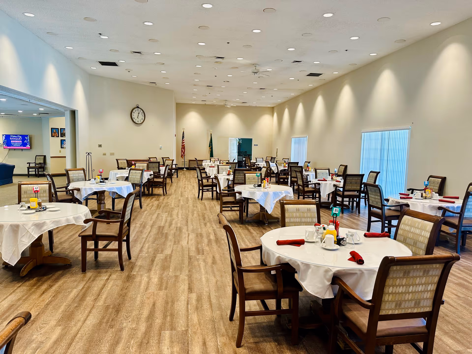 A spacious dining room with multiple round tables covered with white tablecloths, each set with plates, cups, condiments, and red napkins. Wooden chairs with cushioned seats surround the tables. The room has light-colored walls, a wooden floor, ceiling lights, and two windows with vertical blinds. An American flag and another flag are visible at the far end of the room, along with a clock on the wall and a television screen in an adjacent room.