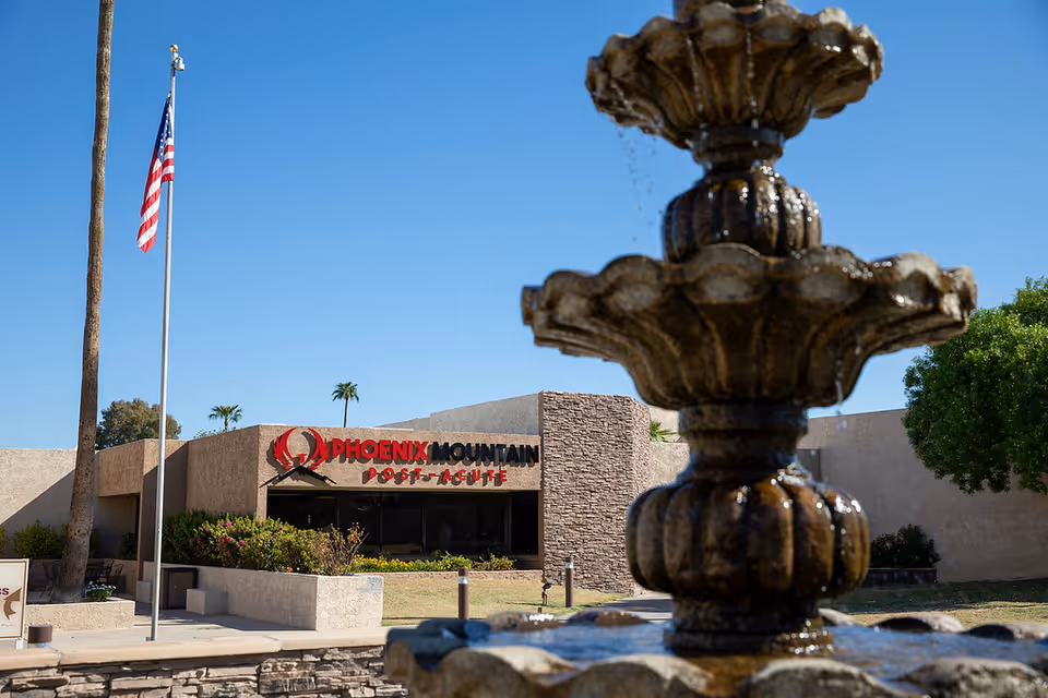 Outdoor view of the Phoenix Mountain Post-Acute facility with a stone fountain in the foreground, an American flag on a flagpole, and clear blue sky.