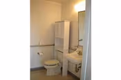 Small senior-living bathroom with a toilet, pedestal sink and mirror, and a tall white storage cabinet.