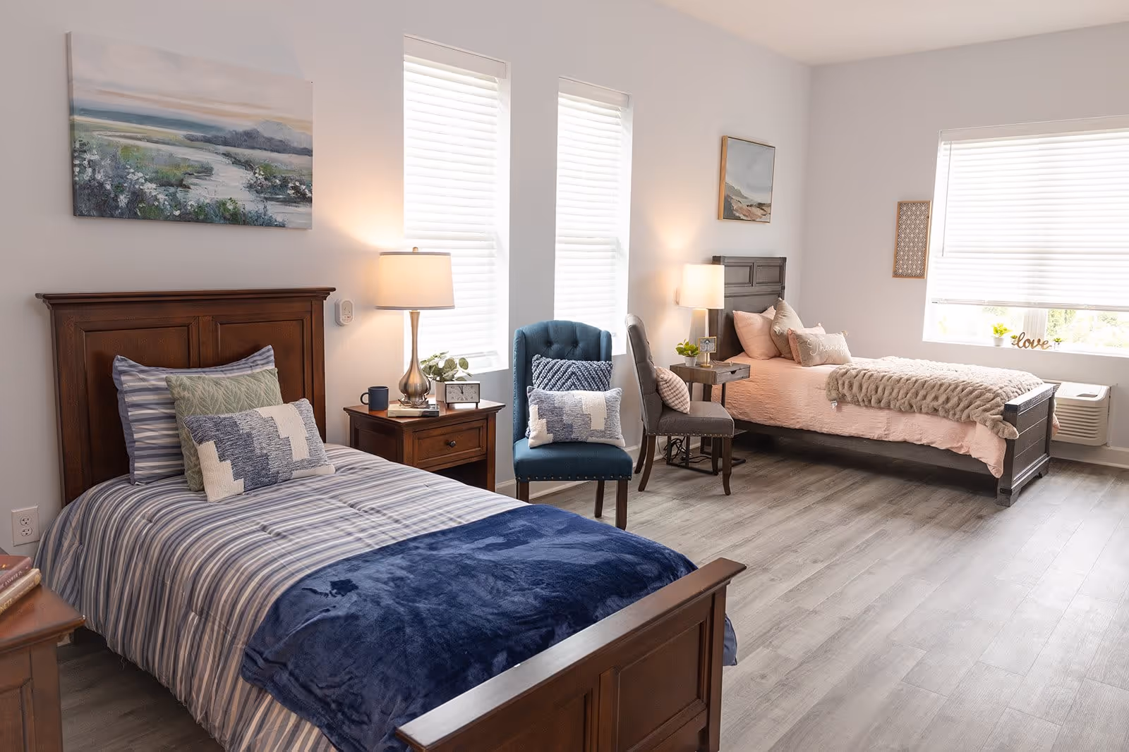 A bright and spacious bedroom with two single wooden beds, one with blue striped bedding and the other with pink bedding. Each bed has multiple pillows and a bedside table with a lamp. There are two windows with white blinds letting in natural light, a blue upholstered chair with a decorative pillow, and framed artwork on the walls. The floor is a light wood laminate.