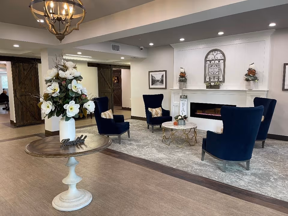 A cozy and elegant common area with four navy blue armchairs arranged around a round coffee table on a gray patterned rug. Behind the seating area is a white fireplace decorated with autumn-themed floral arrangements and a decorative wall piece. In the foreground, there is a round wooden table with a white vase holding large white flowers and a small decorative sign that says 'Thankful'. The room features wooden barn-style sliding doors and a chandelier hanging from the ceiling.