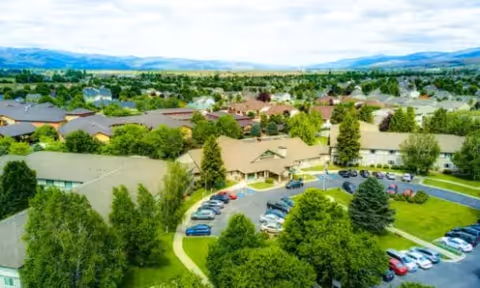 Aerial view of a senior living campus with multiple low-rise buildings, tree-lined grounds, parking lots, and distant mountains.