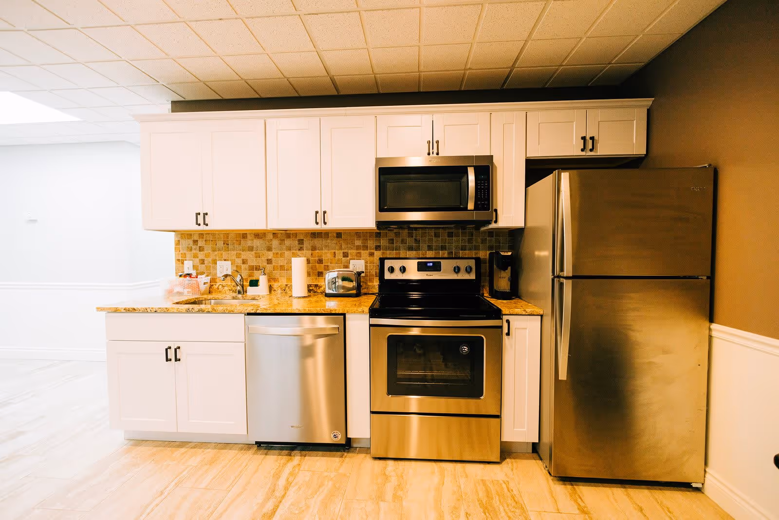 A modern kitchen with white cabinets, a stainless steel refrigerator, oven, microwave, and dishwasher. The countertop is granite with a tiled backsplash, and there is a paper towel holder, toaster, and coffee maker on the counter.