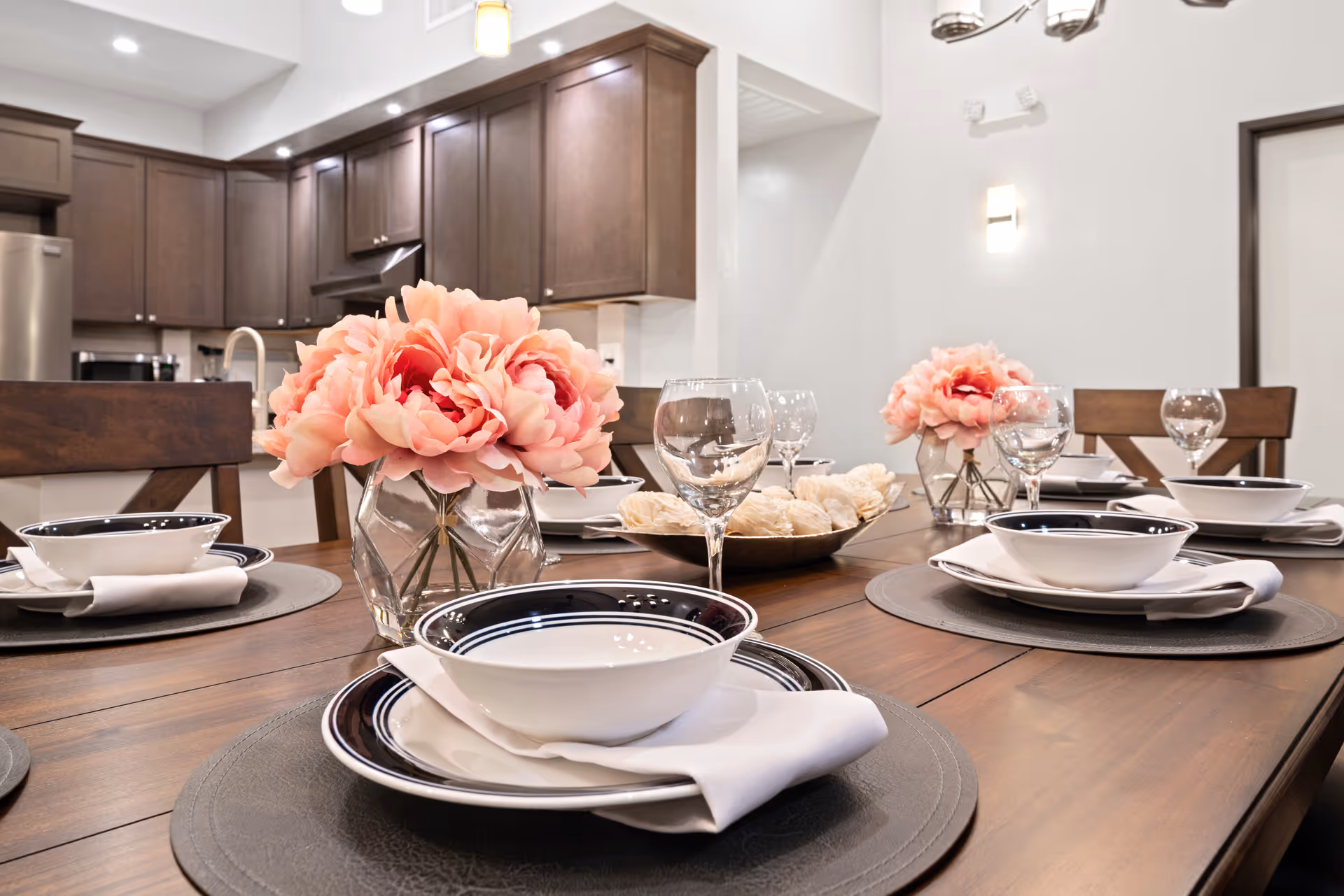 A dining table set for a meal with white bowls, plates, napkins, and wine glasses. Two vases with pink flowers are placed on the table. In the background, there is a kitchen with dark wooden cabinets and stainless steel appliances.