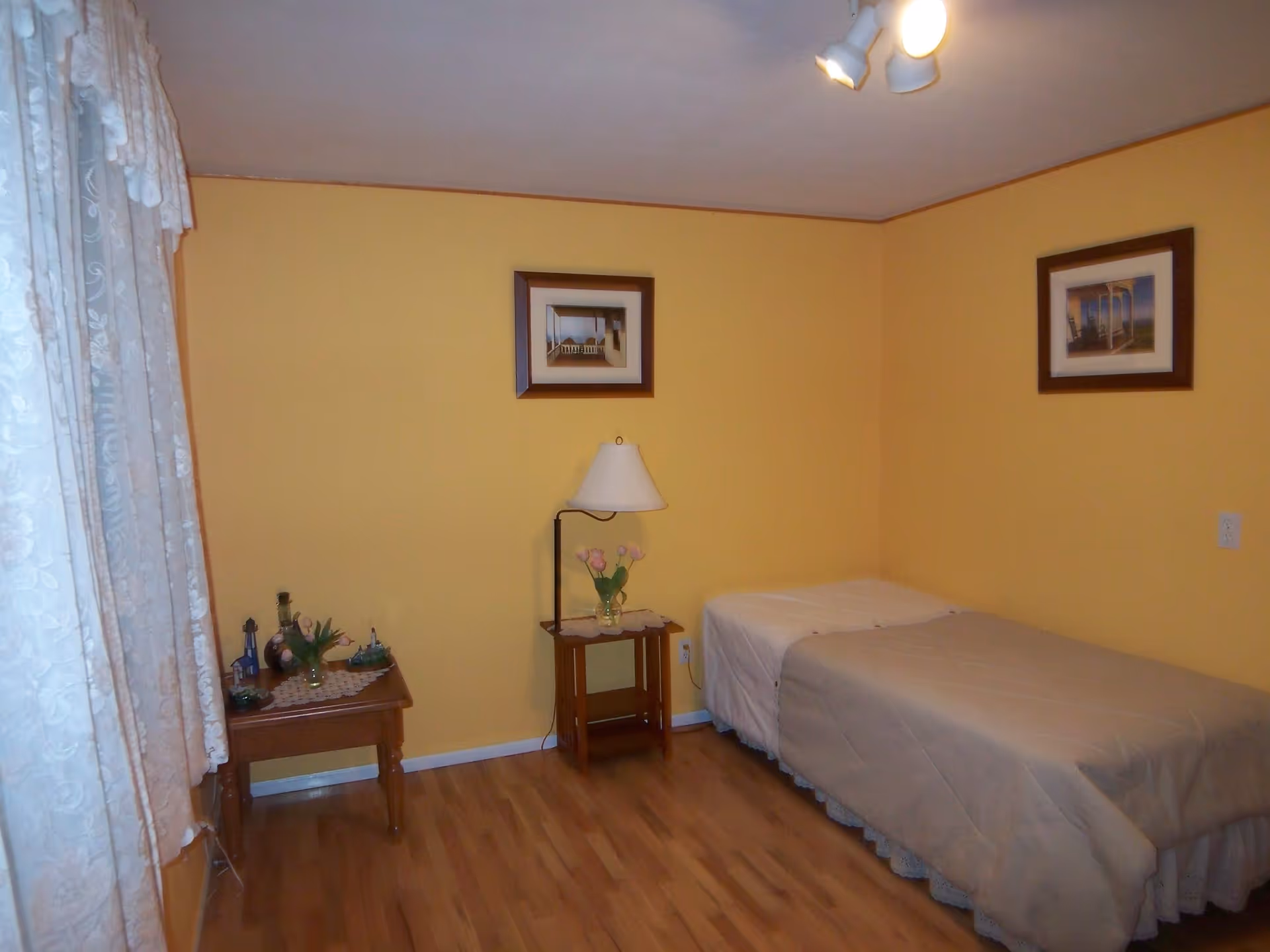 A small bedroom with yellow walls and wooden flooring. There is a single bed with a beige quilt on the right side, a wooden nightstand with a lamp and a vase of pink tulips next to the bed. On the left side, there is a wooden table with decorative items and a vase with flowers. Two framed pictures hang on the walls, and a window with white lace curtains is on the left.