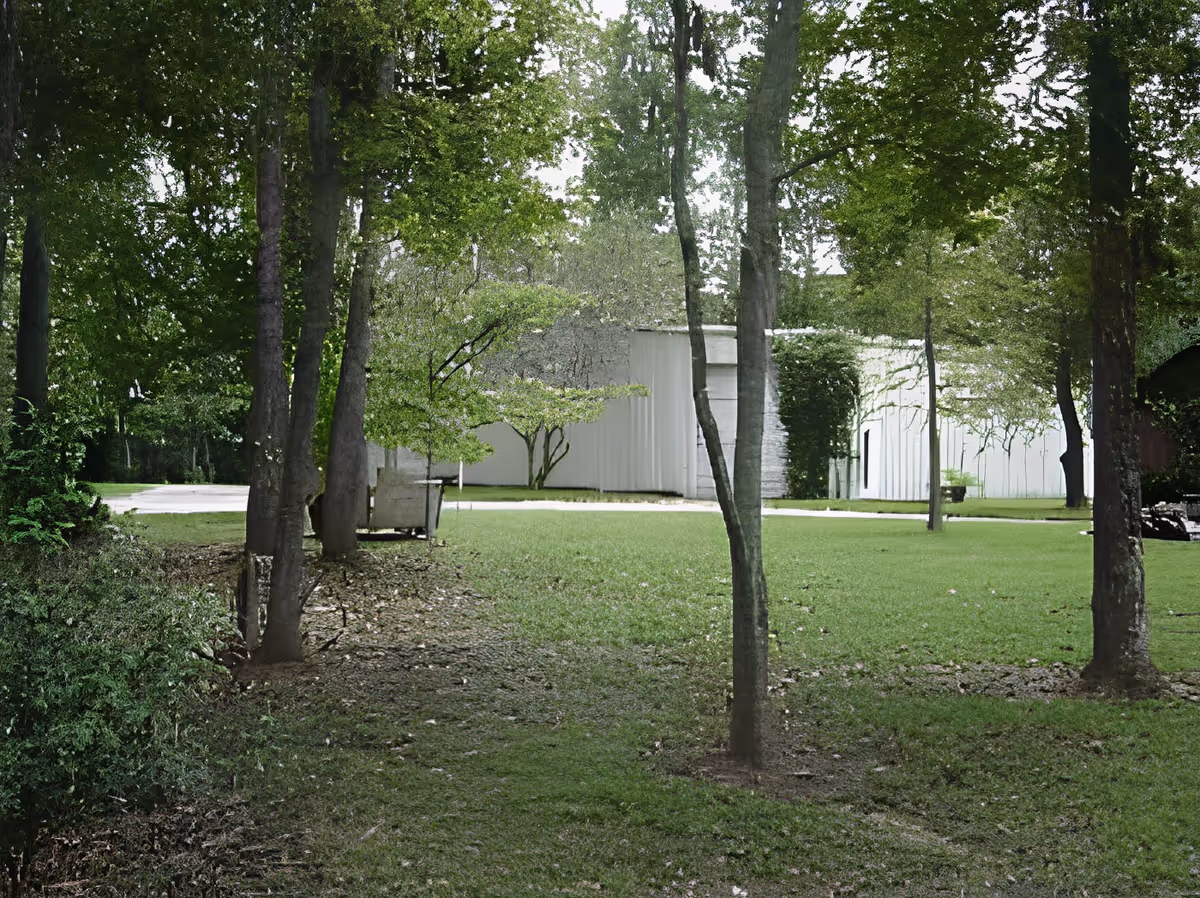 A grassy outdoor area with several trees and bushes, leading to a white building partially covered with ivy in the background.