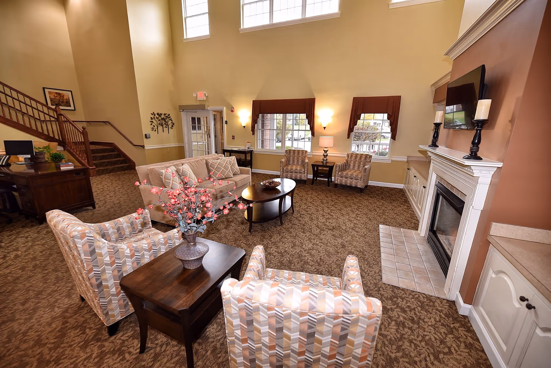 A cozy senior living facility common area with beige walls and carpeted floor. The room features a beige sofa, patterned armchairs, a wooden coffee table with a decorative bowl, and a side table with a vase of pink flowers. There is a fireplace with candles on the mantel and a flat-screen TV mounted above it. Large windows with brown valances let in natural light. A staircase with wooden railings is visible in the background along with a desk and computer.