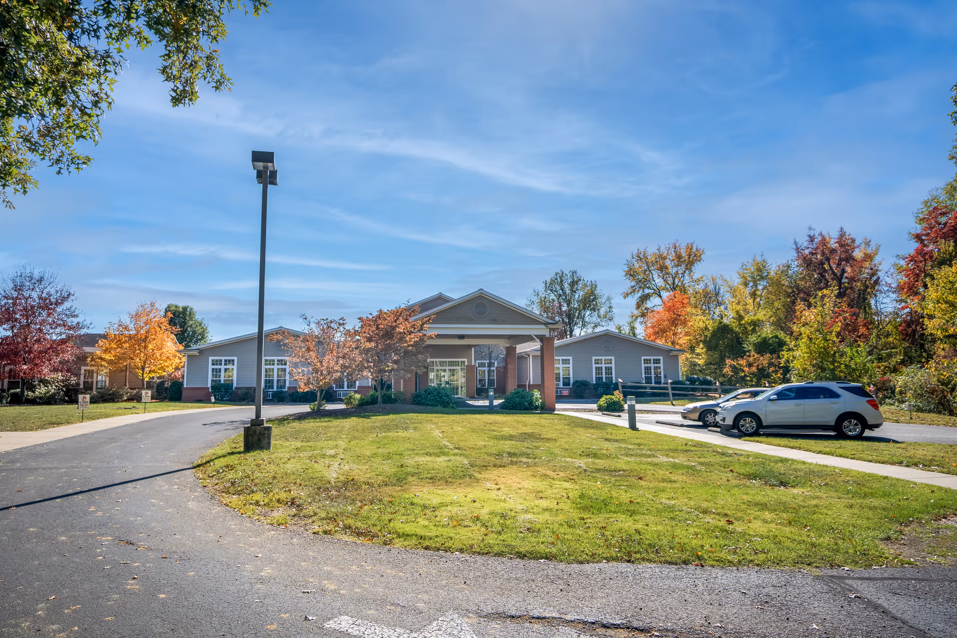 Exterior view of River Bend Nursing & Rehabilitation facility with a driveway, green lawn, several parked cars, and trees with autumn foliage under a clear blue sky.