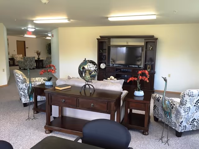 A well-lit living room area in an assisted living facility featuring a dark wood entertainment center with a flat-screen TV, a beige sofa, two floral-patterned armchairs, a wooden console table with a decorative globe and books, and two side tables each holding a vase with red flowers. The room has light-colored walls and carpeted flooring.