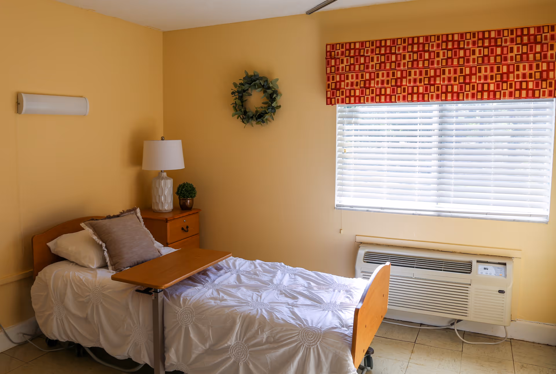 A small bedroom with a single bed covered in a white textured bedspread and two pillows. Next to the bed is a wooden nightstand with a white lamp and a small potted plant. A green wreath hangs on the yellow wall above the nightstand. A window with white blinds and a red patterned valance is on the right side, with an air conditioning unit installed below it.
