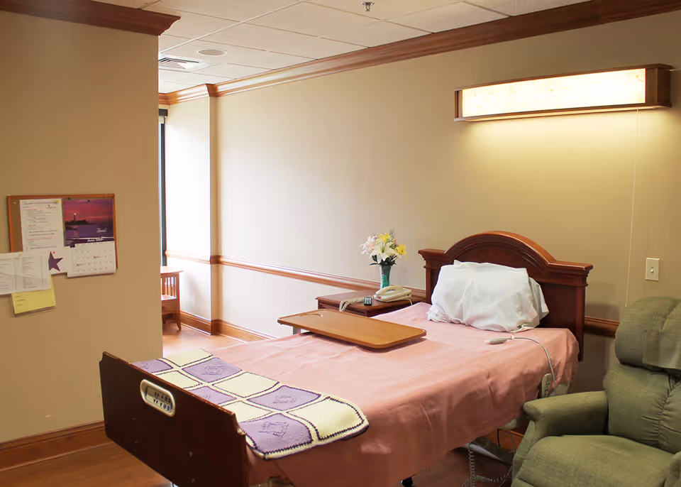 A senior living facility bedroom with a single bed covered in a pink sheet and a purple and white knitted blanket at the foot. The bed has a wooden headboard and a tray table on top. Next to the bed is a wooden nightstand with a vase of flowers, a telephone, and a remote control. A cushioned green recliner chair is positioned beside the bed. The room has beige walls with wooden trim and a large light fixture above the bed. A bulletin board with papers and a calendar is mounted on the wall near a window with natural light coming in.