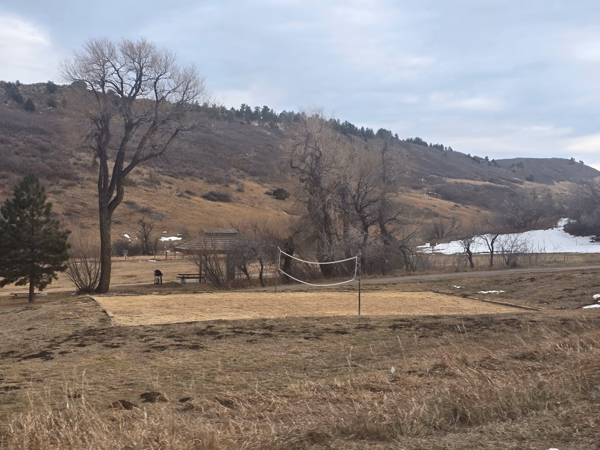 Outdoor scene featuring a sandy volleyball court with a net, surrounded by dry grass and leafless trees. In the background, there are hills with sparse vegetation and patches of snow under a cloudy sky.