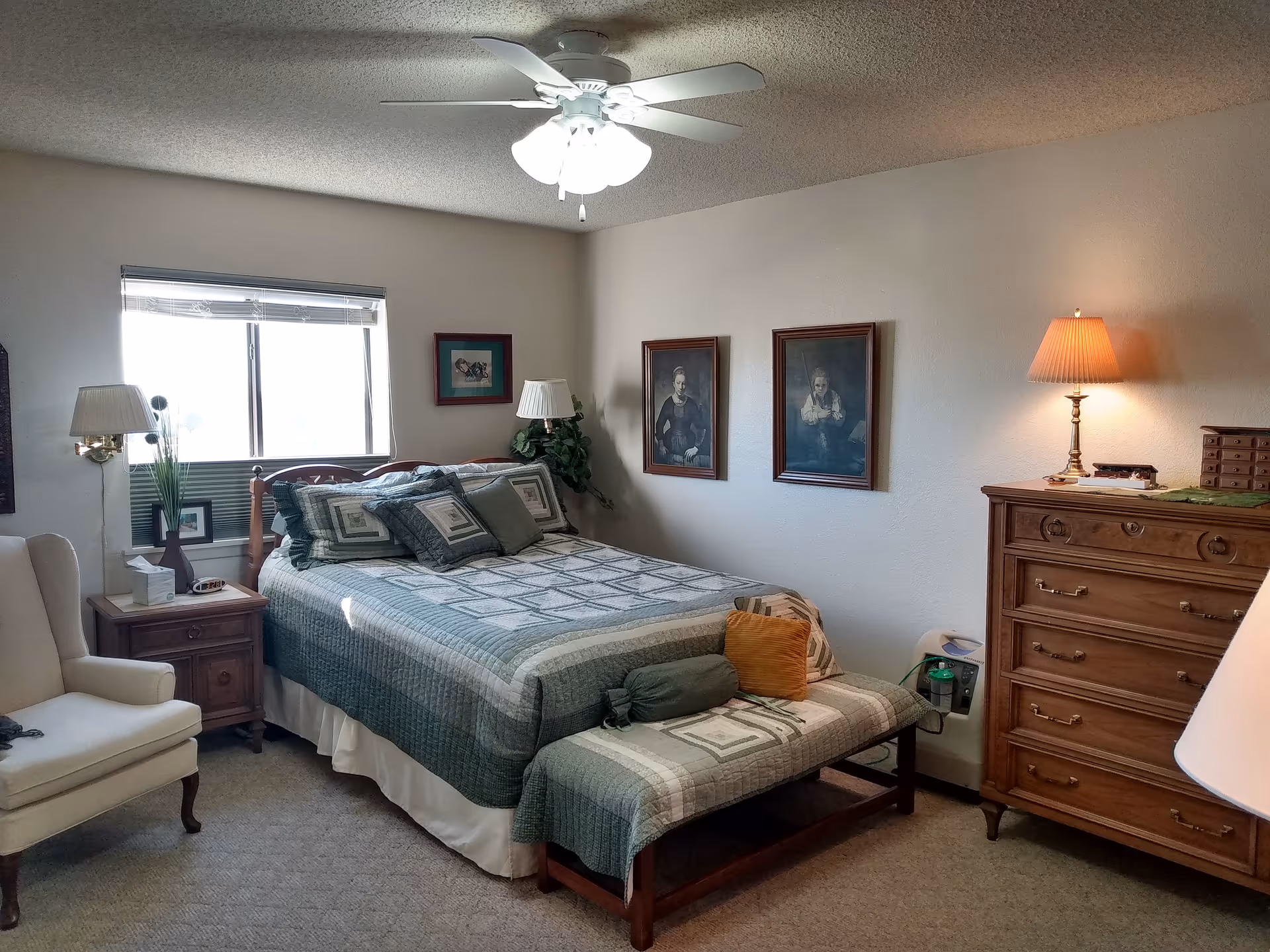 A neatly furnished bedroom with a quilted bed, pillows, nightstands, dresser, armchair, wall art, and a ceiling fan.