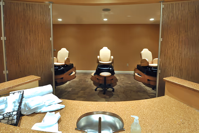Spa pedicure room with three beige pedicure chairs viewed from a countertop sink with towels and soap.