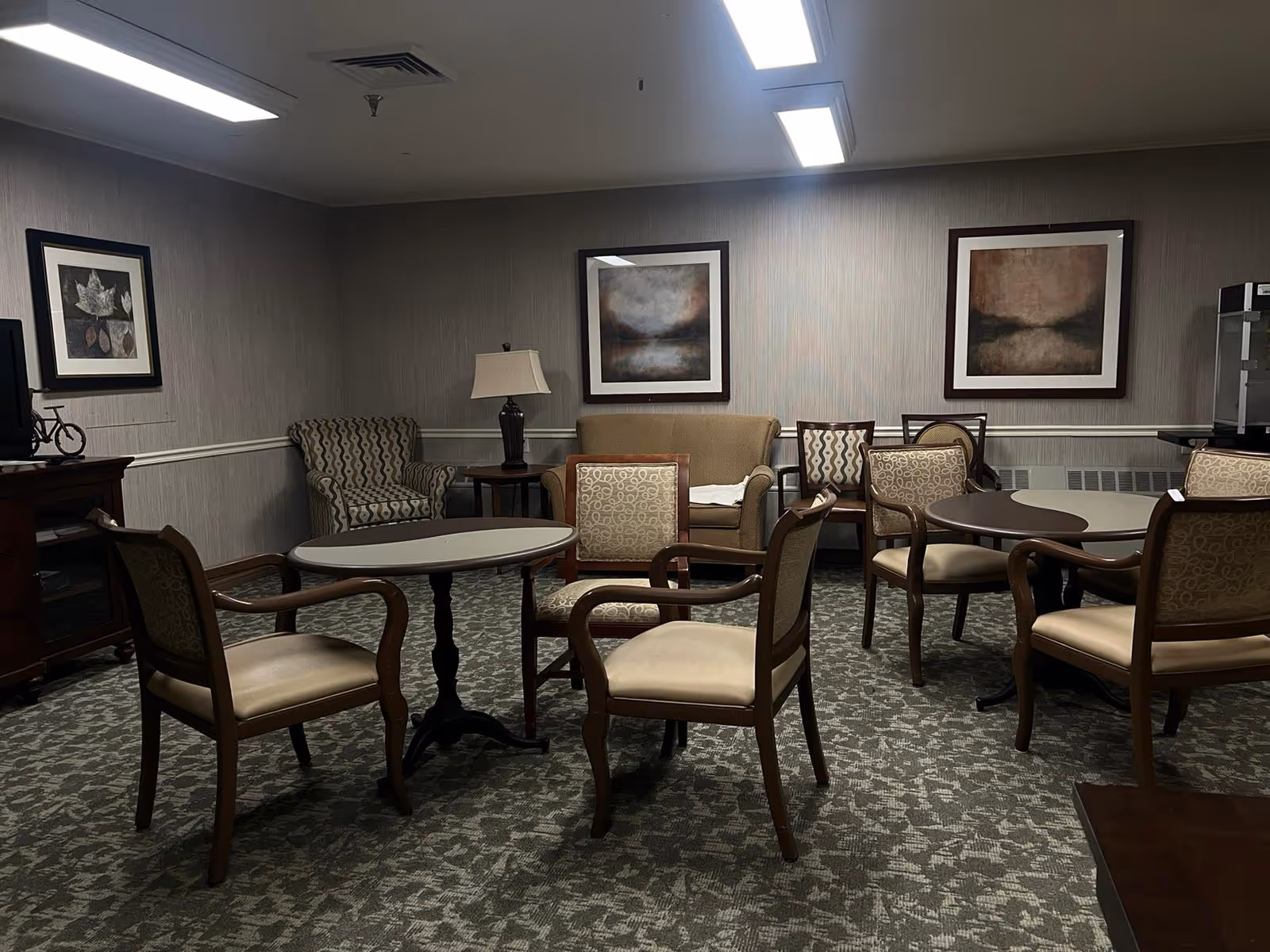 A common area in a senior living facility with several round tables surrounded by wooden chairs with cushioned seats and backs. The room has patterned carpet, neutral-colored walls with three framed abstract paintings, a beige loveseat, an armchair, a side table with a lamp, and a television on a wooden stand.