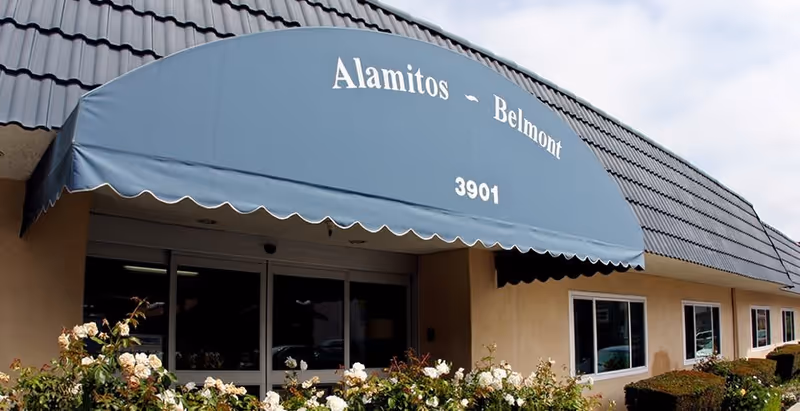 Entrance of Alamitos Belmont Health and Rehabilitation facility with a blue awning displaying the name and address number 3901. The building exterior has beige walls, several windows, and a tiled roof. There are white flowers and green bushes in front of the entrance.