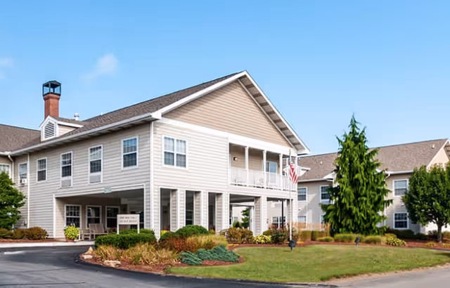 Exterior view of a two-story senior living building with a covered entrance, balconies, landscaped lawn, and an American flag.