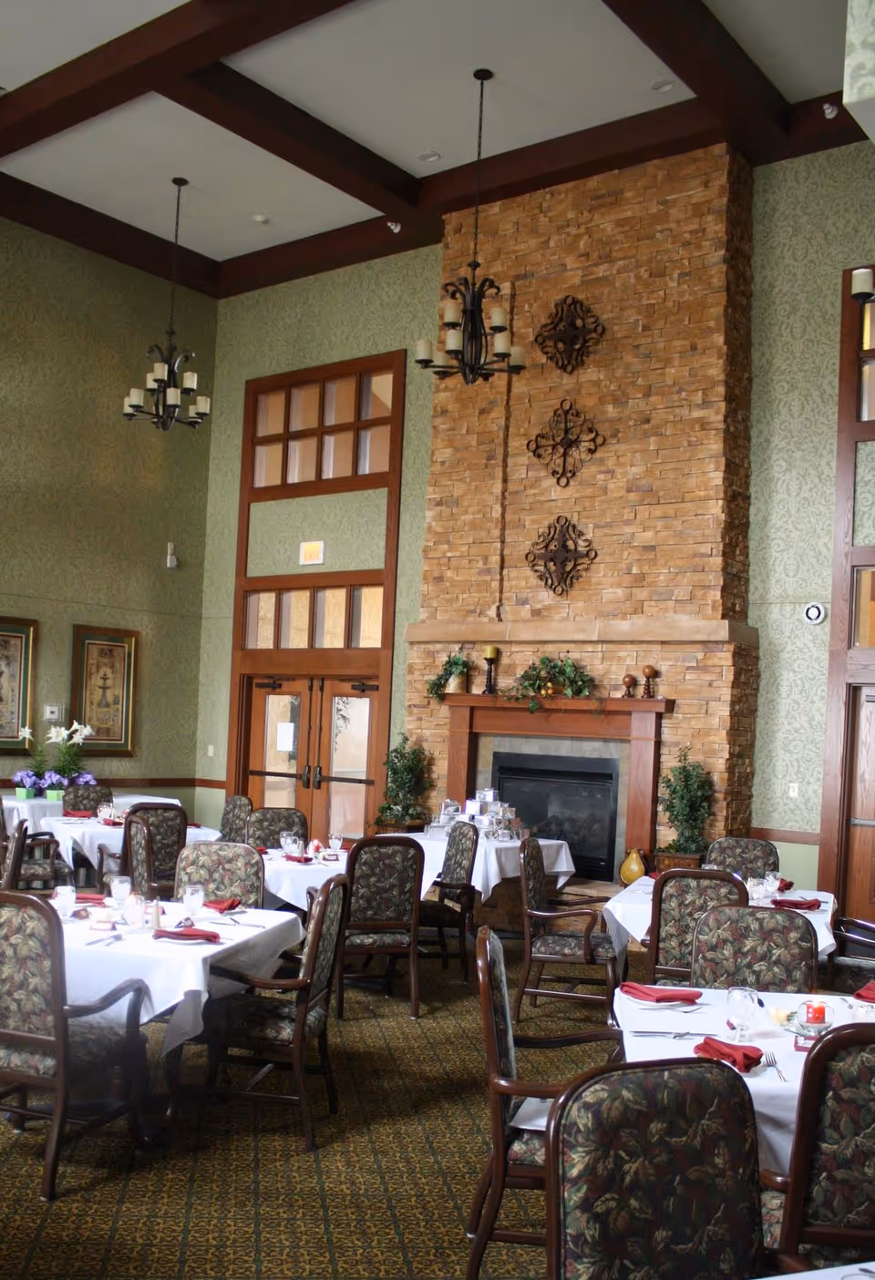 Dining room with multiple tables covered in white tablecloths and set with glasses, plates, and red napkins. The room features a tall stone fireplace decorated with greenery and candle holders, patterned upholstered chairs, green wallpaper, and two chandeliers hanging from a ceiling with dark wooden beams.