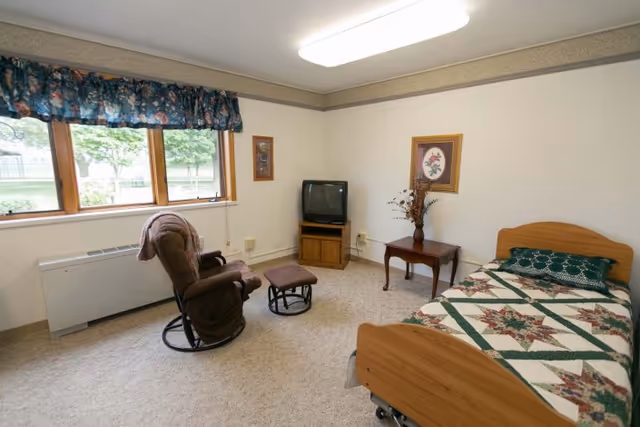A simple, cozy bedroom in a senior living facility with a single bed covered in a patterned quilt, a brown reclining chair with matching footrest, a small wooden side table with a vase of flowers, an old-style television on a wooden stand, and a window with floral curtains letting in natural light.