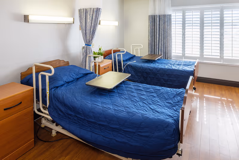 A bright room with two hospital-style beds covered in blue quilts, each with an overbed table. The room has wooden floors, a wooden nightstand between the beds with a plant on it, and large windows with white shutters and blue curtains.