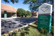 Exterior view of a building with a paved driveway and green landscaping. A sign in the foreground reads 'Living Center at Geneva North' with a heart-shaped logo above the text. Trees and part of the building entrance are visible in the background under a clear blue sky.