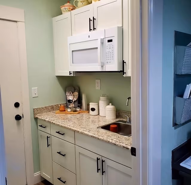 A small kitchen area with light green walls, white cabinets with black handles, a granite countertop, a stainless steel sink with a red cup inside, and a white microwave mounted above the counter. On the countertop, there are several ceramic containers and a wooden tray with kitchen utensils. A white door with black knobs is visible on the left side.